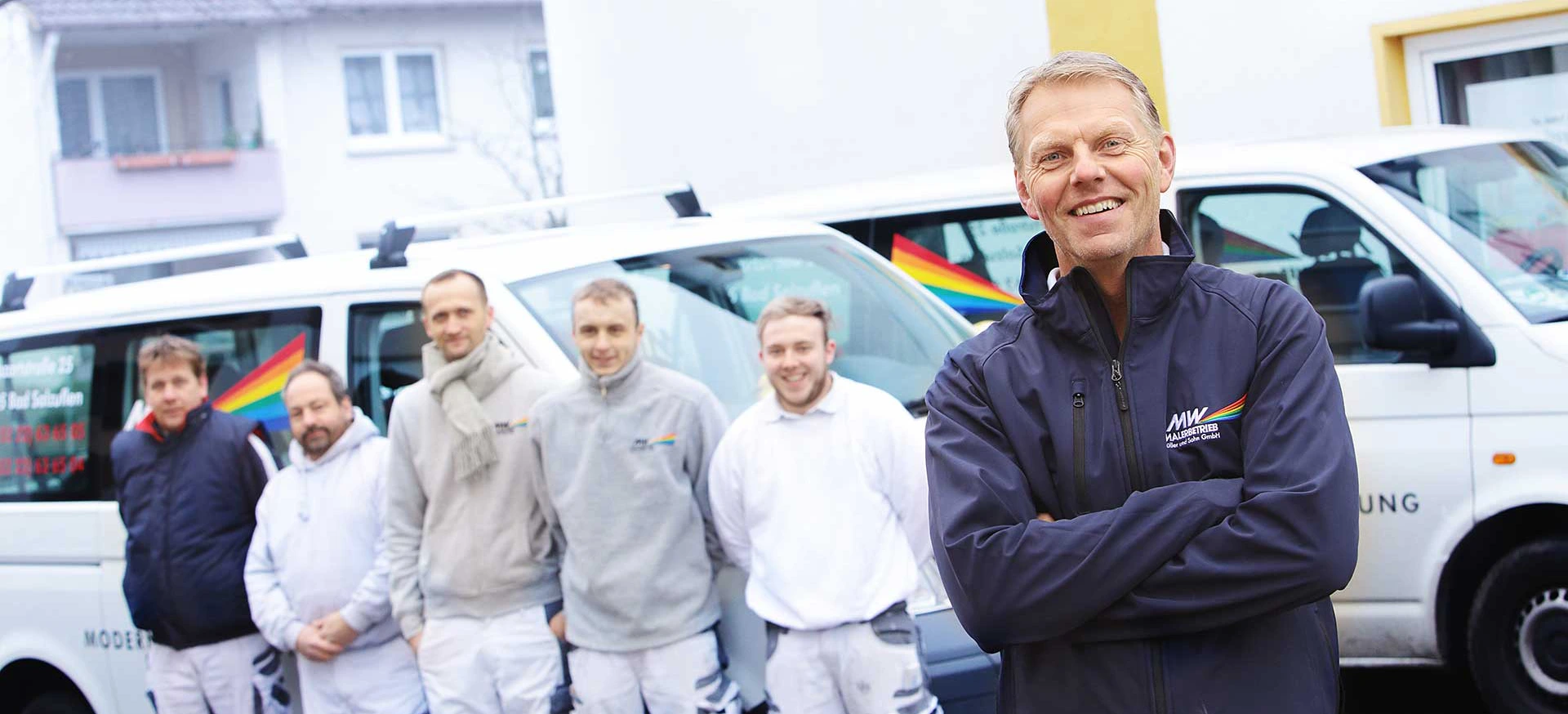 Sechs Mitarbeiter des MW Malerbetriebs Müller & Sohn GmbH lächeln freundlich in die Kamera, während sie in Arbeitskleidung stehen. Im Hintergrund ist das Firmenlogo sichtbar.